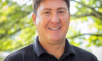 Headshot of Chacon wearing a black collar shirt in front of a natural background.
