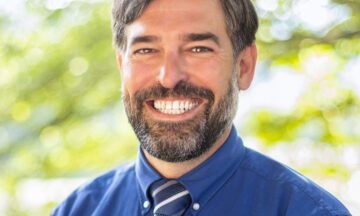 Headshot of Condie wearing a blue button up shirt and a striped tie.