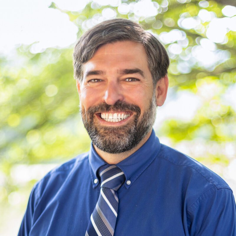 Headshot of Condie wearing a blue button up shirt and a striped tie.