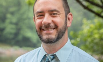Headshot of Dillman with a light blue button up top and a blue and gray tie.