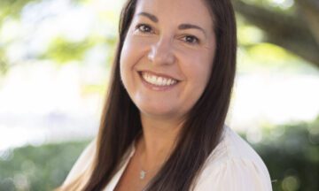 Headshot of Kelly wearing a white blouse before a natural background.
