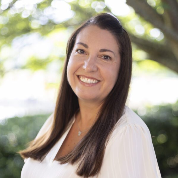 Headshot of Kelly wearing a white blouse before a natural background.