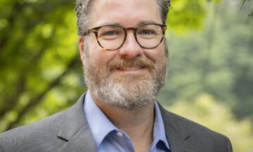 Headshot of Phelan wearing a gray blazer over a powder blue button up shirt.