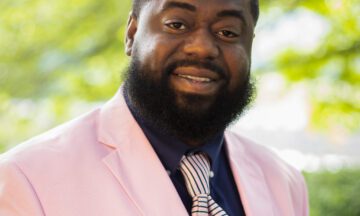Headshot of Clark wearing a pink suit in front of a natural backdrop.
