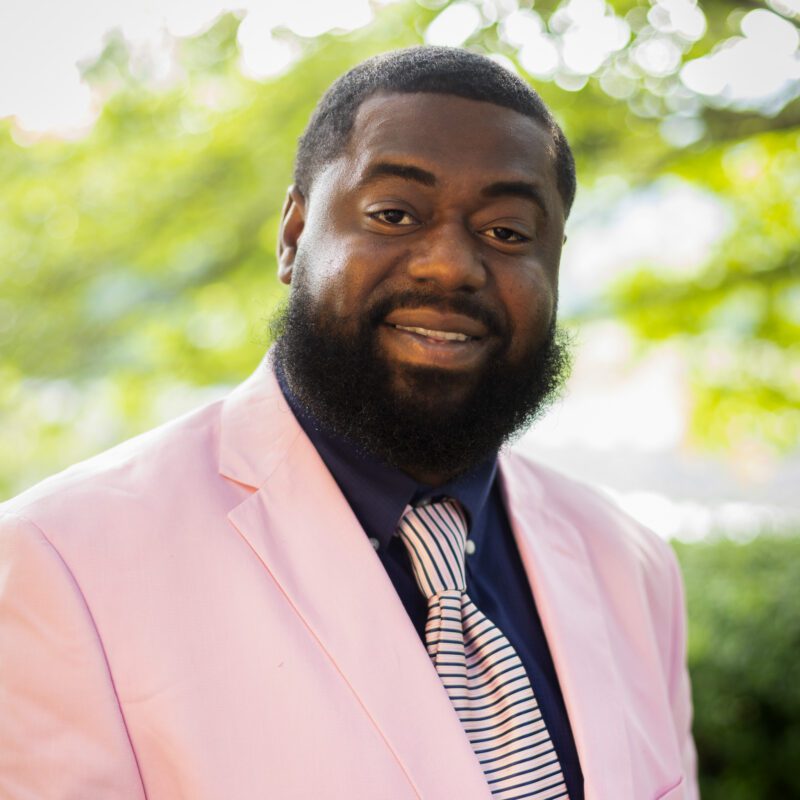 Headshot of Clark wearing a pink suit in front of a natural backdrop.