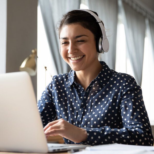 Smiling Indian girl wearing headphones using laptop, looking at screen, happy young female listening to favorite music while working online on project, excited student learning language