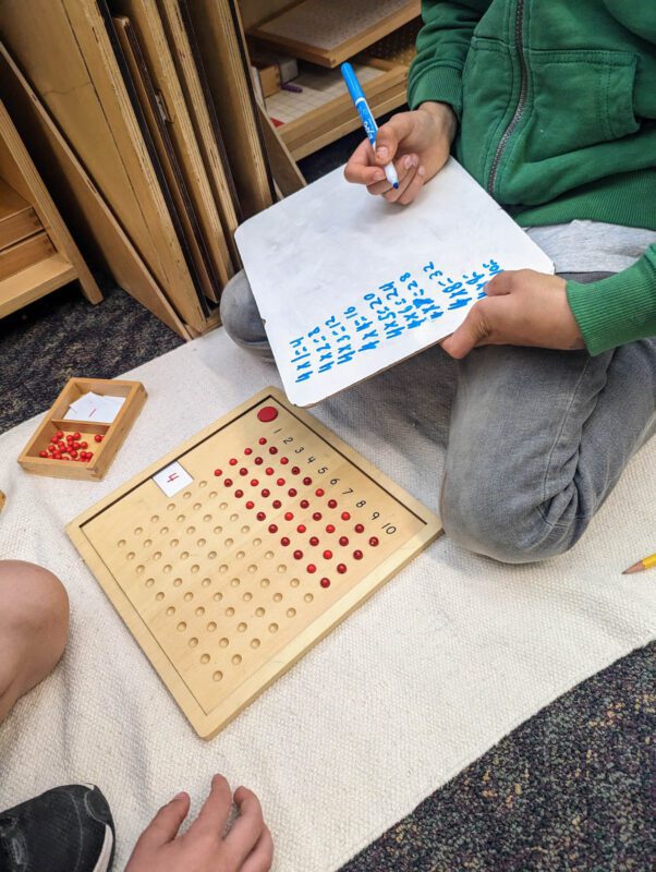 Two students do multiplication using a wooden board and red chips for counting.