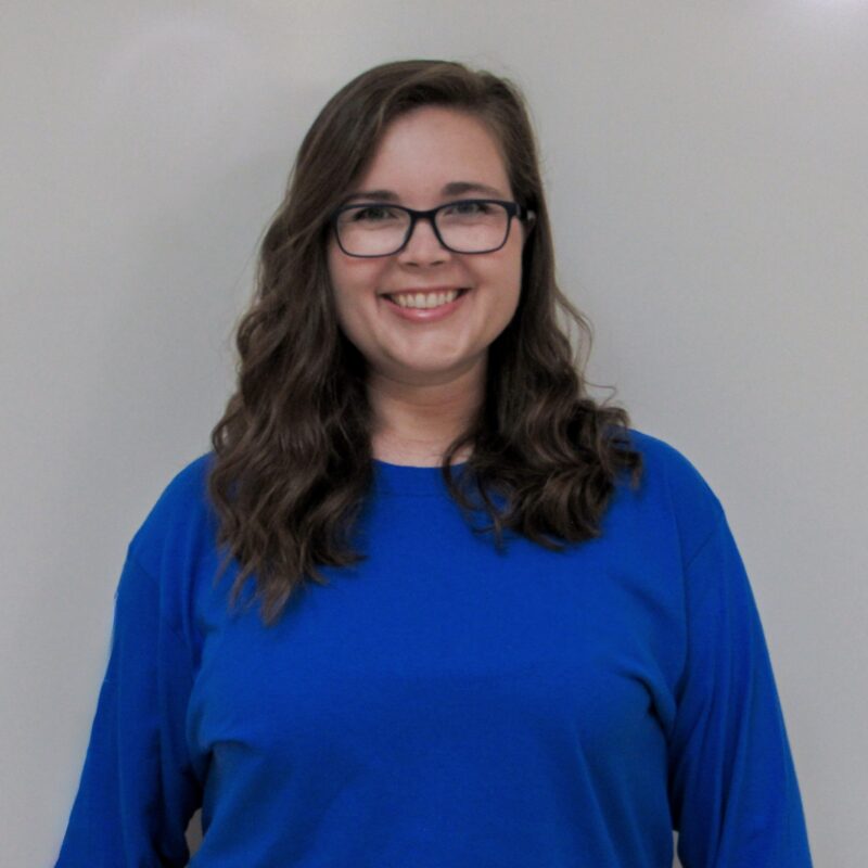 Headshot of Maggie DeCorah wearing a blue blouse standing in front of a gray background.