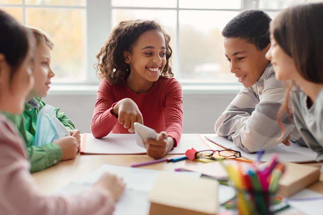 Multiethnic School Children Having Fun Using Smartphone During Break Sitting At Desk In Modern Classroom. African American Schoolgirl Showing New Educational Application On Phone To Classmates